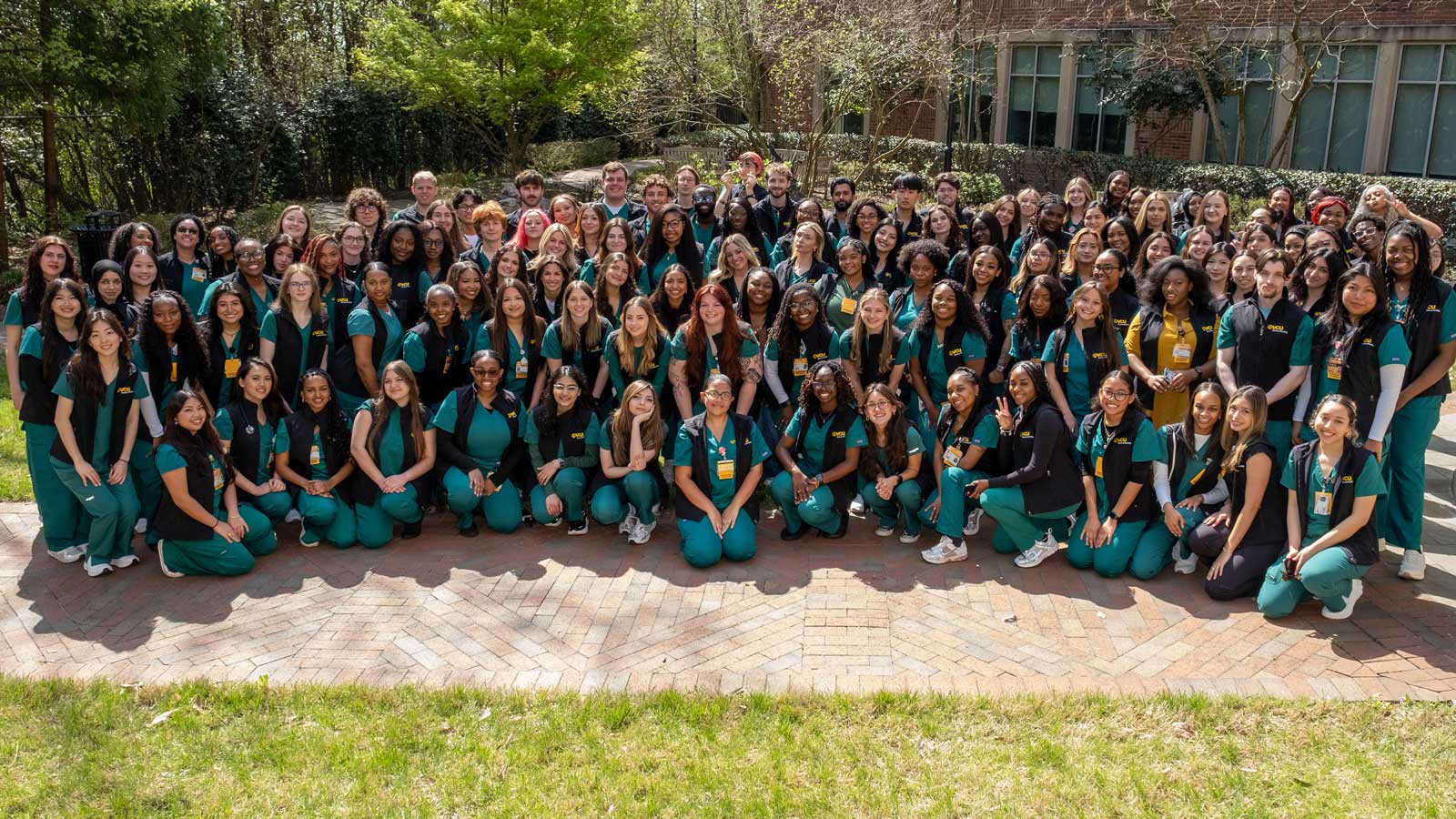 A large group of V.C.U. nursing students in scrubs and vests pose together outdoors on a campus walkway