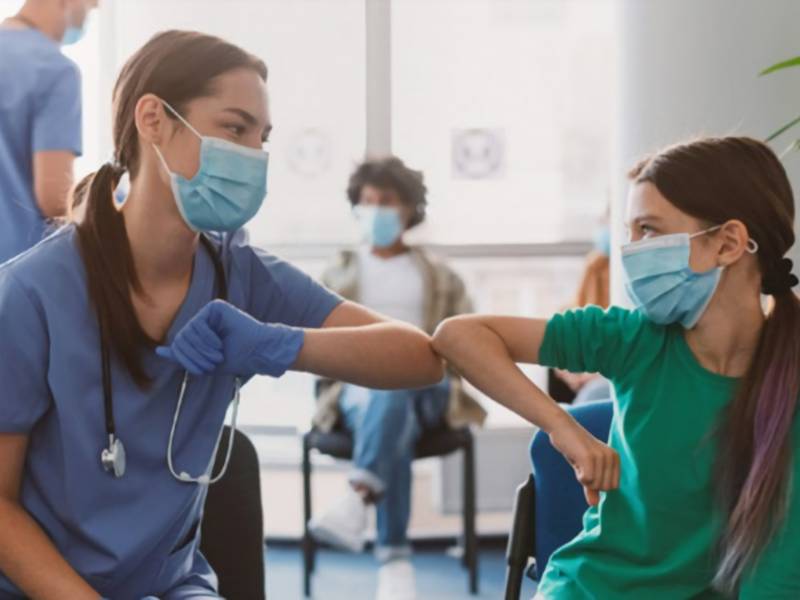 a nurse with a facemask and a child with a facemask bump elbows